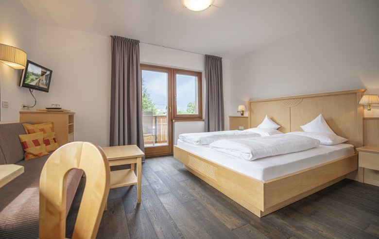 La camera comfort con un letto matrimoniale, un divano e una televisione. Attraverso la porta del balcone si può ammirare la vista diretta sulla natura.