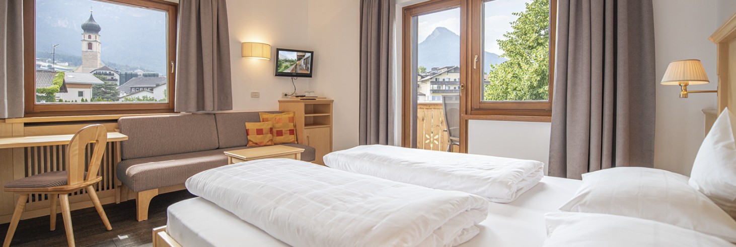 hubertus dolomites a Fiè allo Sciliar | Comfort nella natura: Le nostre camere ai piedi dello Sciliar. La camera con un letto matrimoniale, un divano, una TV e una scrivania. Attraverso le finestre si può ammirare la vista sul villaggio e sulla natura circostante.