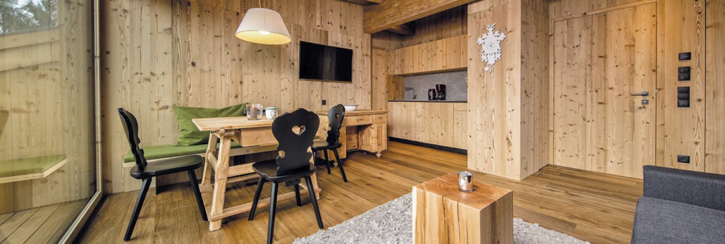 hubertus dolomites a Fiè allo Sciliar | Vivi il comfort e la tradizione nelle nostre camere e appartamenti sull'Alpe di Siusi. L'area soggiorno dell'appartamento. Si vede un tavolo in legno con sedie nere e la cucina sullo sfondo.