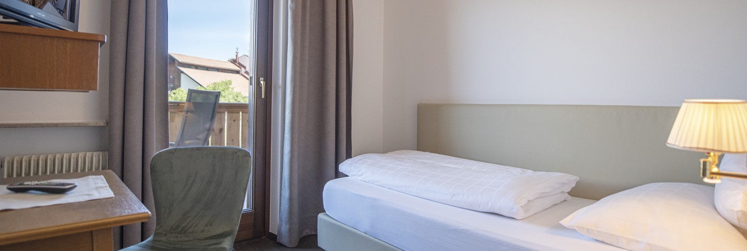 hubertus dolomites a Fiè allo Sciliar | Dormire in armonia con la natura. Una camera singola con letto, scrivania e televisore. È visibile anche una parte del balcone adiacente.