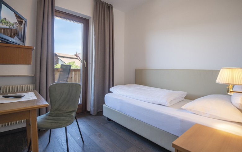 Una camera singola con letto, scrivania e televisore. È visibile anche una parte del balcone adiacente.