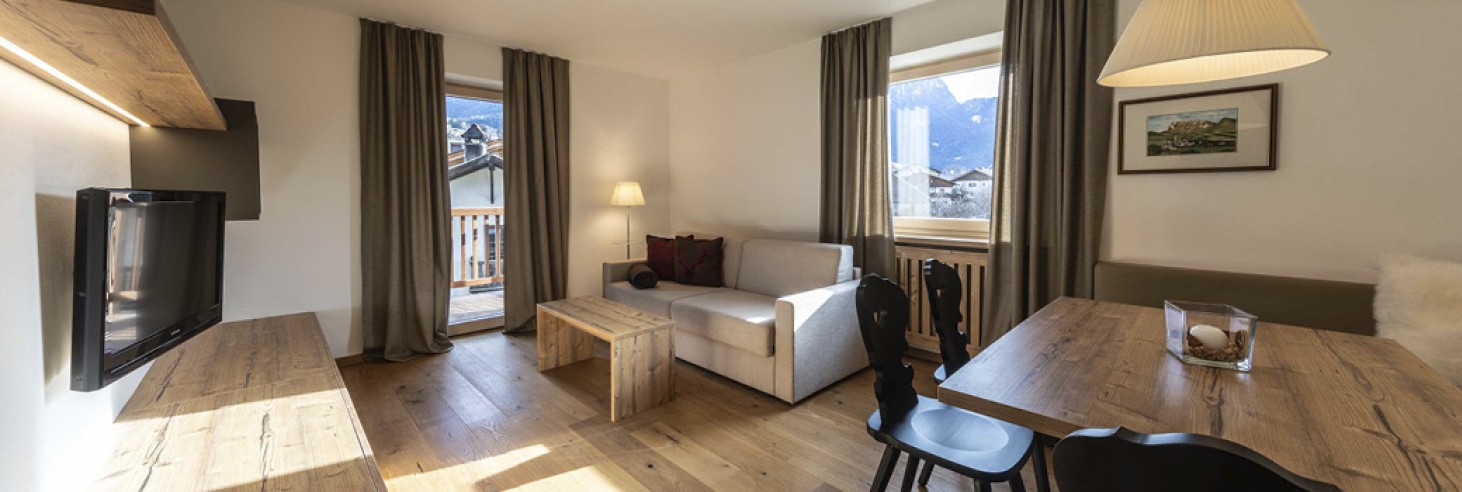 hubertus dolomites a fiè allo sciliar | Una casa nelle Dolomiti: Le nostre sistemazioni sull'Alpe di Siusi. La zona giorno dell'appartamento. Si vedono la TV e il divano, così come il tavolo da pranzo con sedie nere.