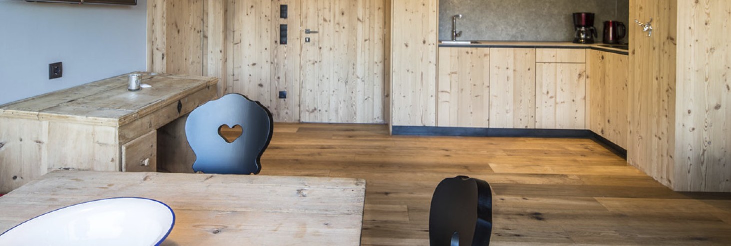 hubertus dolomites a Fiè allo Sciliar | Tranquillità e comfort nelle nostre camere sull'Alpe di Siusi. La zona giorno dell'appartamento vacanze è arredato completamente in legno, con in primo piano il tavolo in legno e le sedie nere. Sullo sfondo si vedono la zona cucina e la TV.