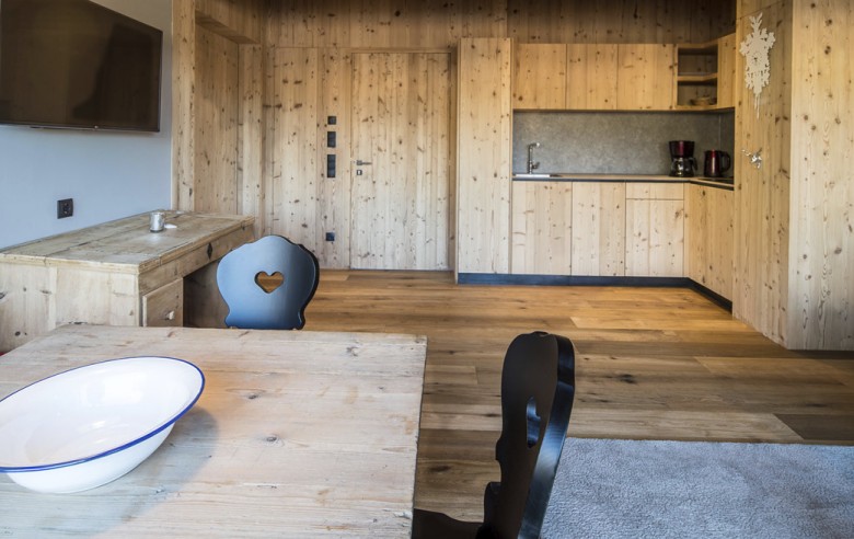 La zona giorno dell'appartamento vacanze è arredato completamente in legno, con in primo piano il tavolo in legno e le sedie nere. Sullo sfondo si vedono la zona cucina e la TV.