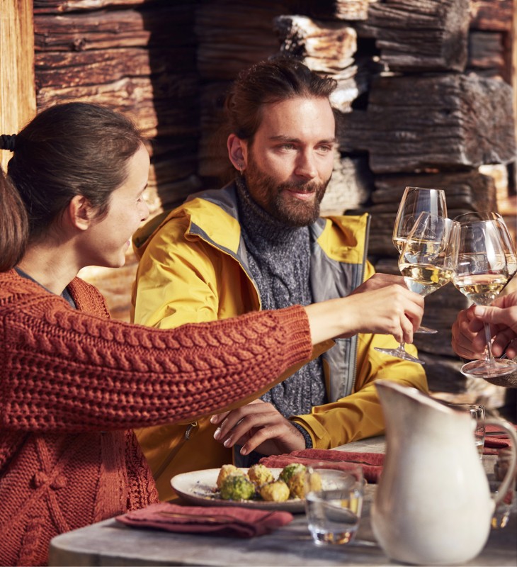 hubertus dolomites Fiè allo Sciliar | Scoperte culinarie in Alto Adige: ristoranti e piatti locali Una coppia in una baita di montagna. Sta pranzando e bevendo un bicchiere di vino bianco altoatesino.