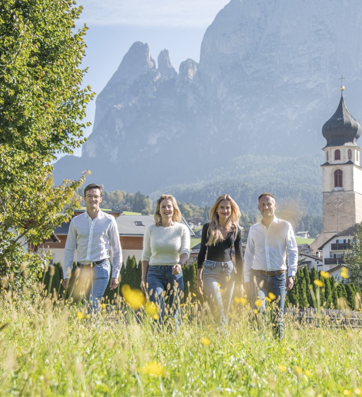 hubertus dolomites Fié allo Sciliar | Posizione perfetta nel cuore del paese, direttamente ai piedi dello Sciliar La famiglia Profanter in mezzo a un prato con lo Schlern e il campanile della chiesa sullo sfondo.