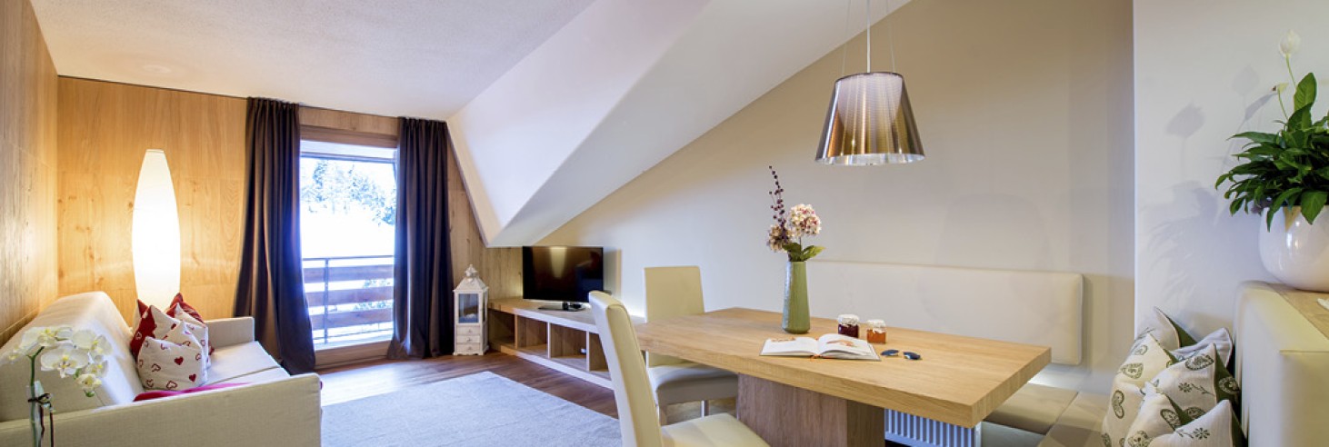 hubertus dolomites a fiè allo sciliar | Appartamenti sull'Alpe di Siusi: Dove natura e comfort si fondono. La zona giorno dell'appartamento. Si vedono un tavolo da pranzo, una TV e un divano. Inoltre, si intravede anche il balcone e la vista sulla natura.