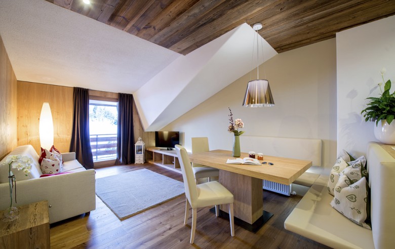 La zona giorno dell'appartamento. Si vedono un tavolo da pranzo, una TV e un divano. Inoltre, si intravede anche il balcone e la vista sulla natura.