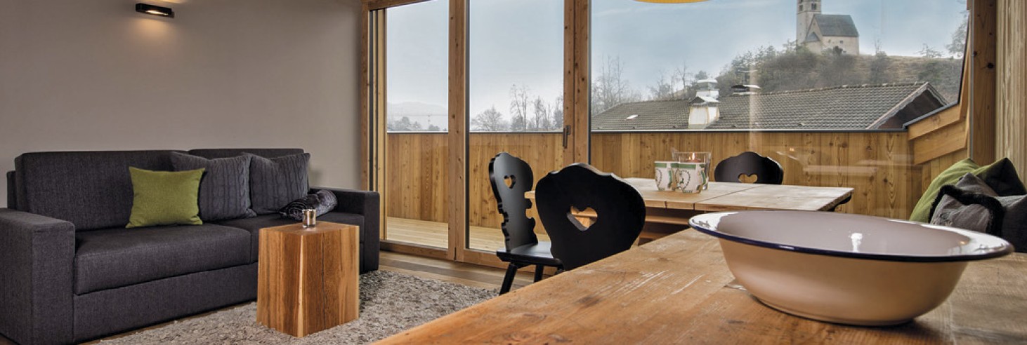 hubertus dolomites a Fiè allo Sciliar | Il tuo rifugio nelle montagne dell'Alto Adige. La zona giorno della camera. Si vedono un divano, un tavolo in legno con sedie nere e il balcone con vista.